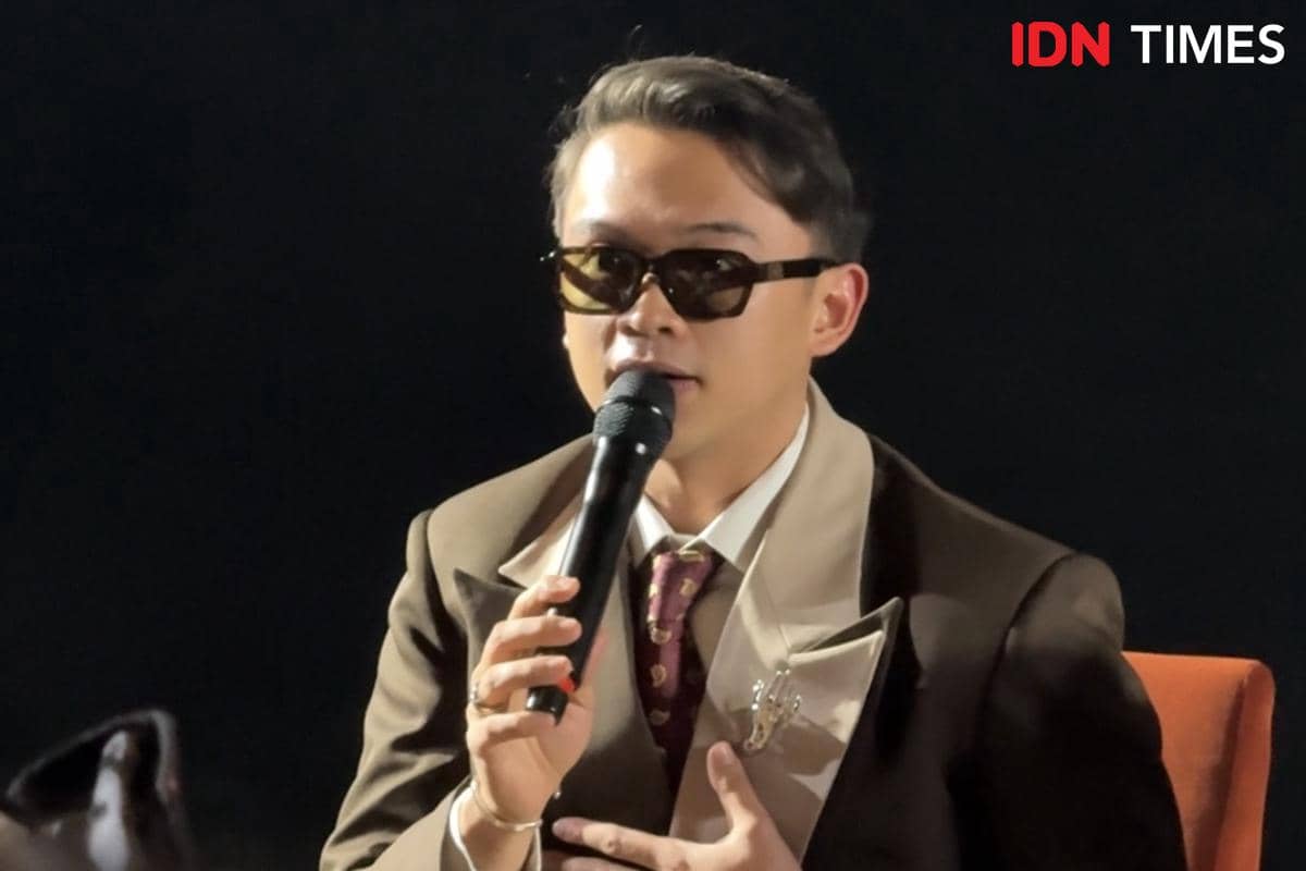 Seorang pria mengenakan setelan jas cokelat dan kacamata hitam sedang berbicara memegang mikrofon dalam konferensi pers film Monster Pabrik Rambut.