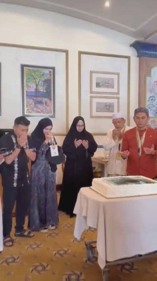 Beberapa orang berpakaian rapi berdiri berdoa di depan kue besar di atas meja yang tertutup kain putih di sebuah ruangan berhias lukisan.