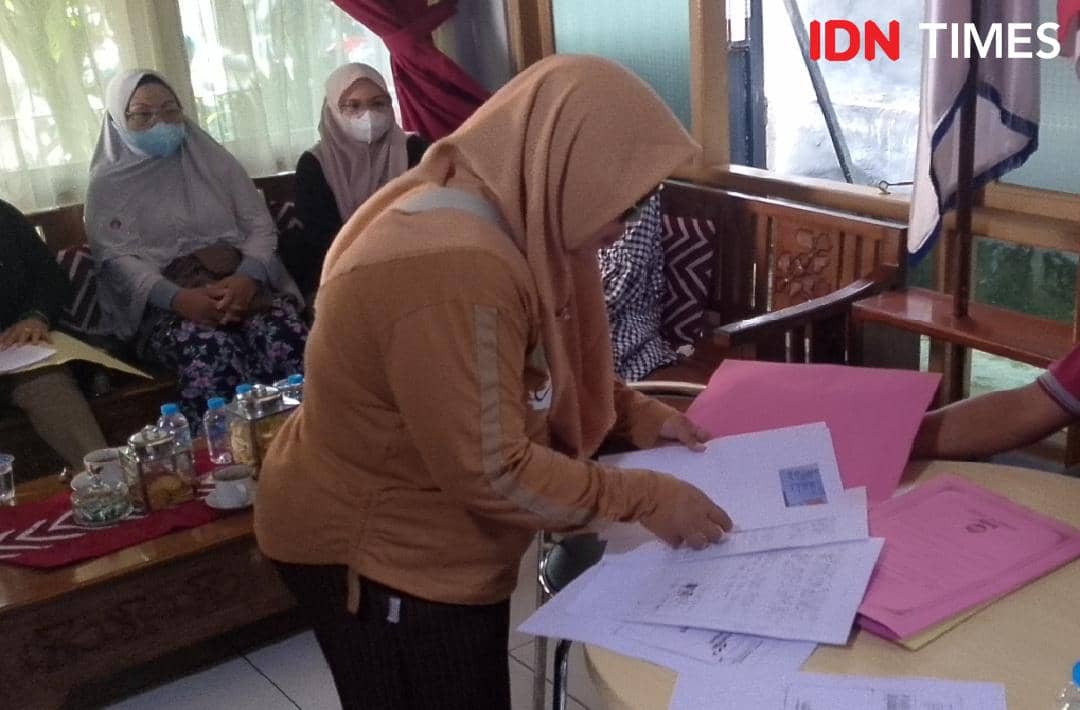 Seorang perempuan berhijab memeriksa dokumen di meja, sementara beberapa orang lain duduk di belakangnya di sebuah ruangan kantor.
