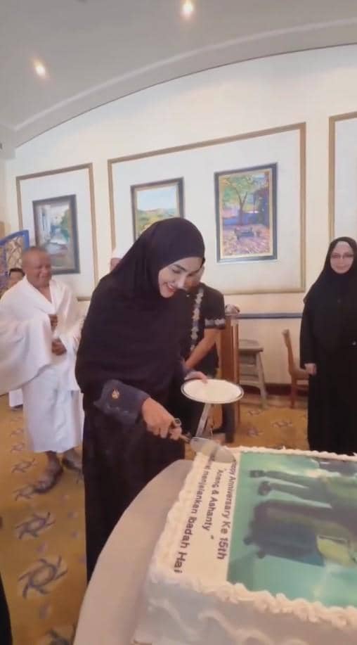 Seorang wanita berhijab hitam tersenyum sambil memotong kue besar bertuliskan ucapan ulang tahun pernikahan di sebuah ruangan.