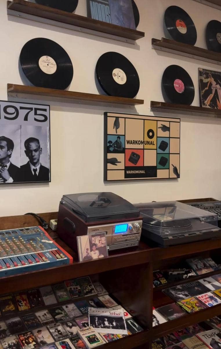 Interior kafe Warkomunal di Bandar Lampung dengan dekorasi piringan hitam, poster musik, dan koleksi kaset serta pemutar vinyl retro.
