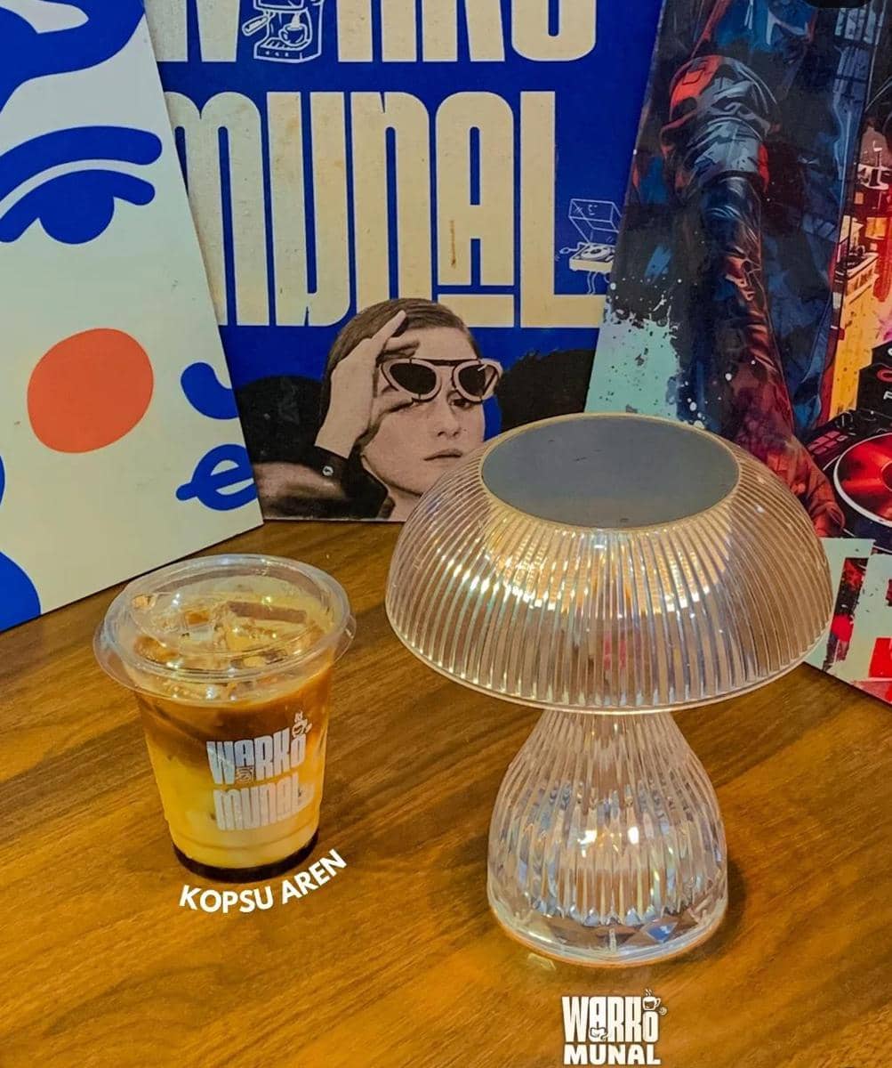 Segelas kopi susu gula aren di atas meja kayu dengan lampu meja kaca dan latar dekorasi artistik di kafe Warkomunal Bandar Lampung.