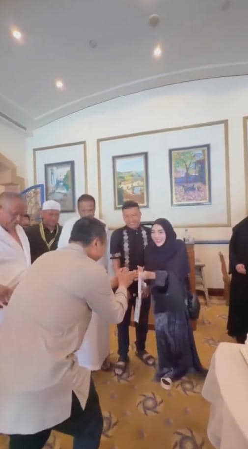 Sepasang suami istri berpakaian sopan berwarna hitam dan abu-abu berjabat tangan dengan seseorang di sebuah ruangan bersama beberapa orang lainnya.
