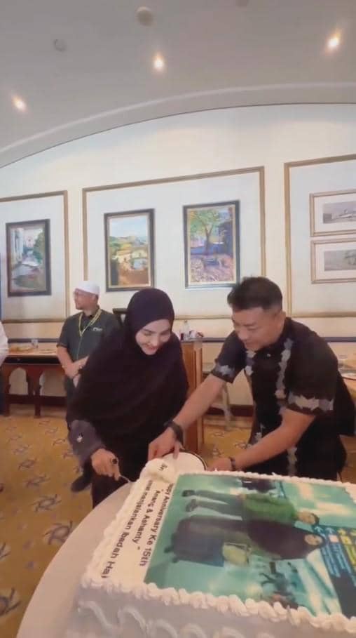 Sepasang suami istri berpakaian hitam memotong kue besar bertema perayaan di sebuah ruangan dengan dekorasi lukisan di dinding.