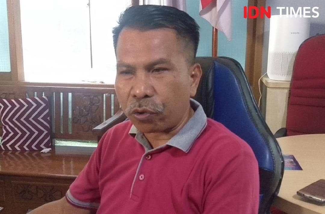 Seorang pria mengenakan kaus polo merah duduk di kursi kantor biru di dalam ruangan dengan meja dan kursi di sekitarnya.