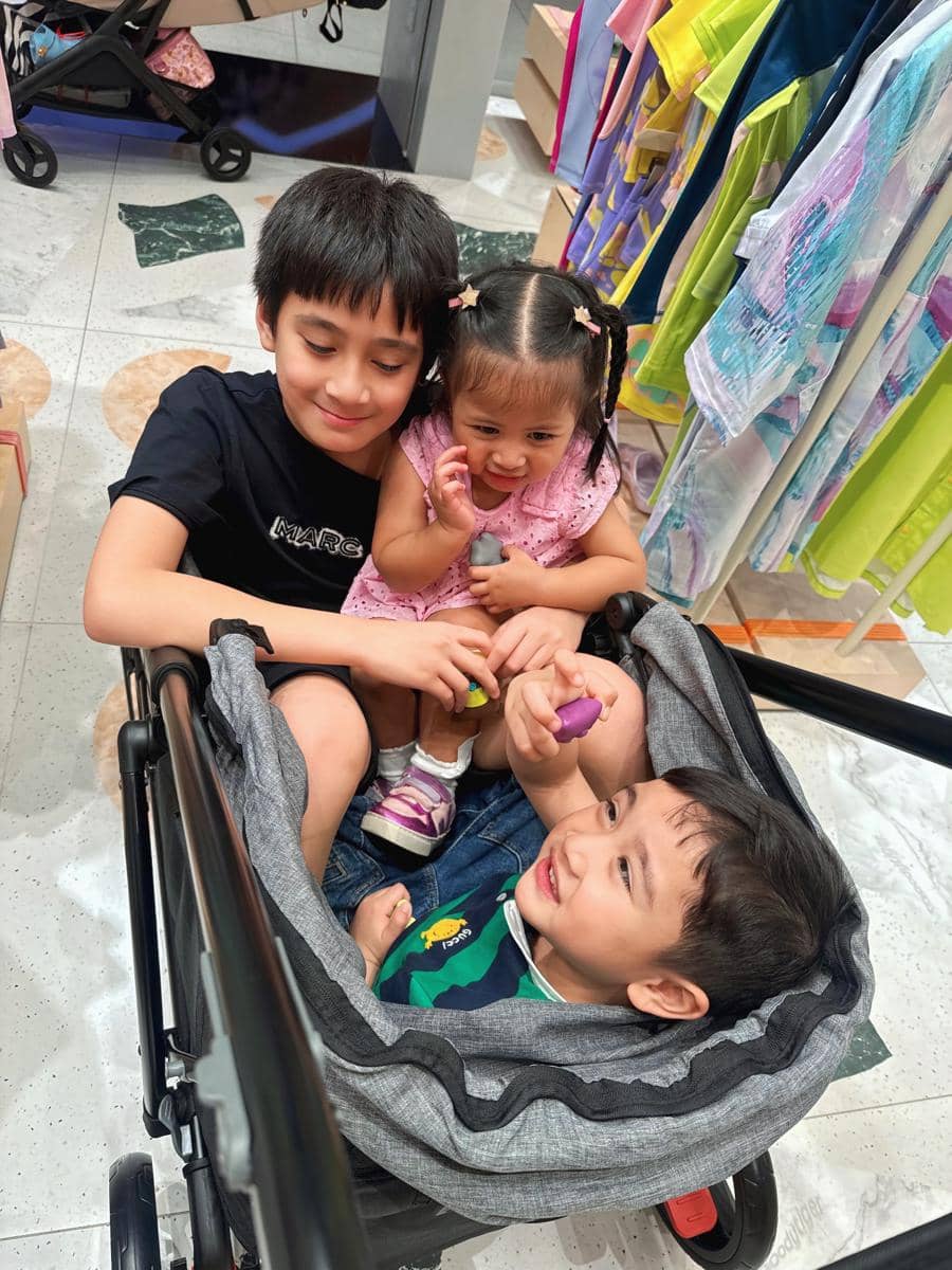 Tiga anak kecil tersenyum dan bermain bersama di dalam stroller saat berlibur di Dubai, dikelilingi pakaian berwarna cerah.
