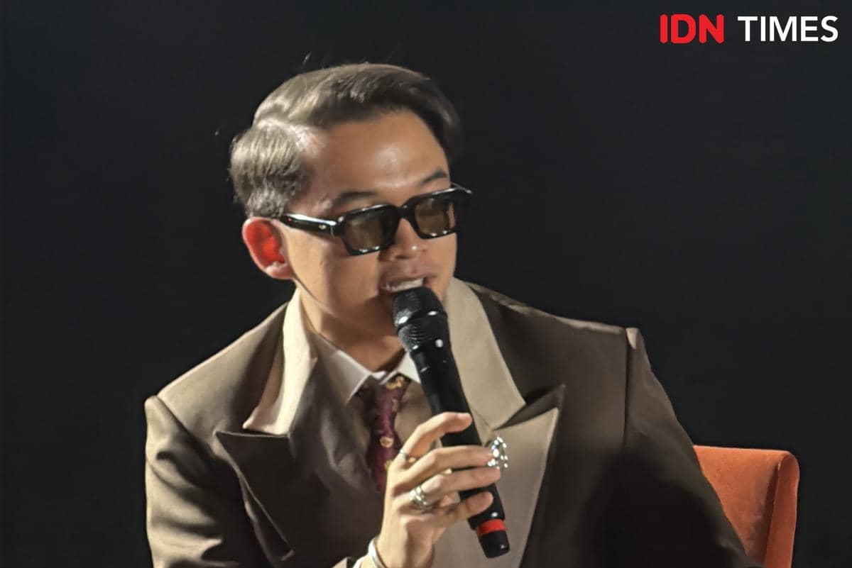 Seorang pria mengenakan jas cokelat dan kacamata hitam berbicara sambil memegang mikrofon dalam konferensi pers film Monster Pabrik Rambut.