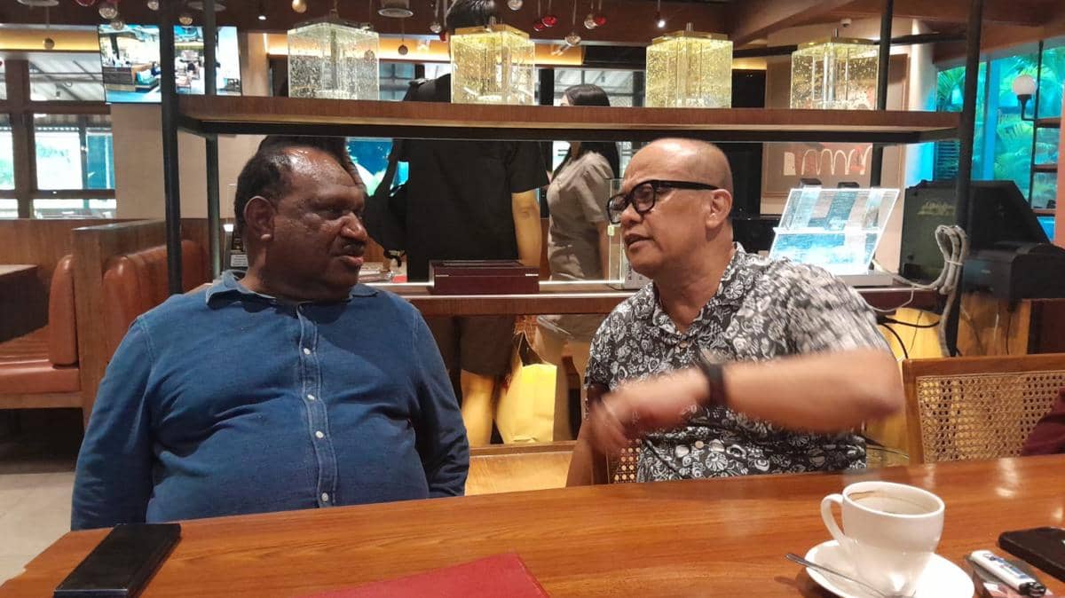 Dua pria sedang berbincang di sebuah kafe dengan suasana santai, salah satunya mengenakan kemeja biru dan yang lain berkemeja motif.