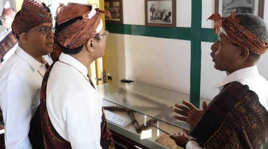 Tiga pria mengenakan pakaian adat dan ikat kepala tradisional sedang berbincang di dalam ruangan dengan pajangan foto dan benda bersejarah.