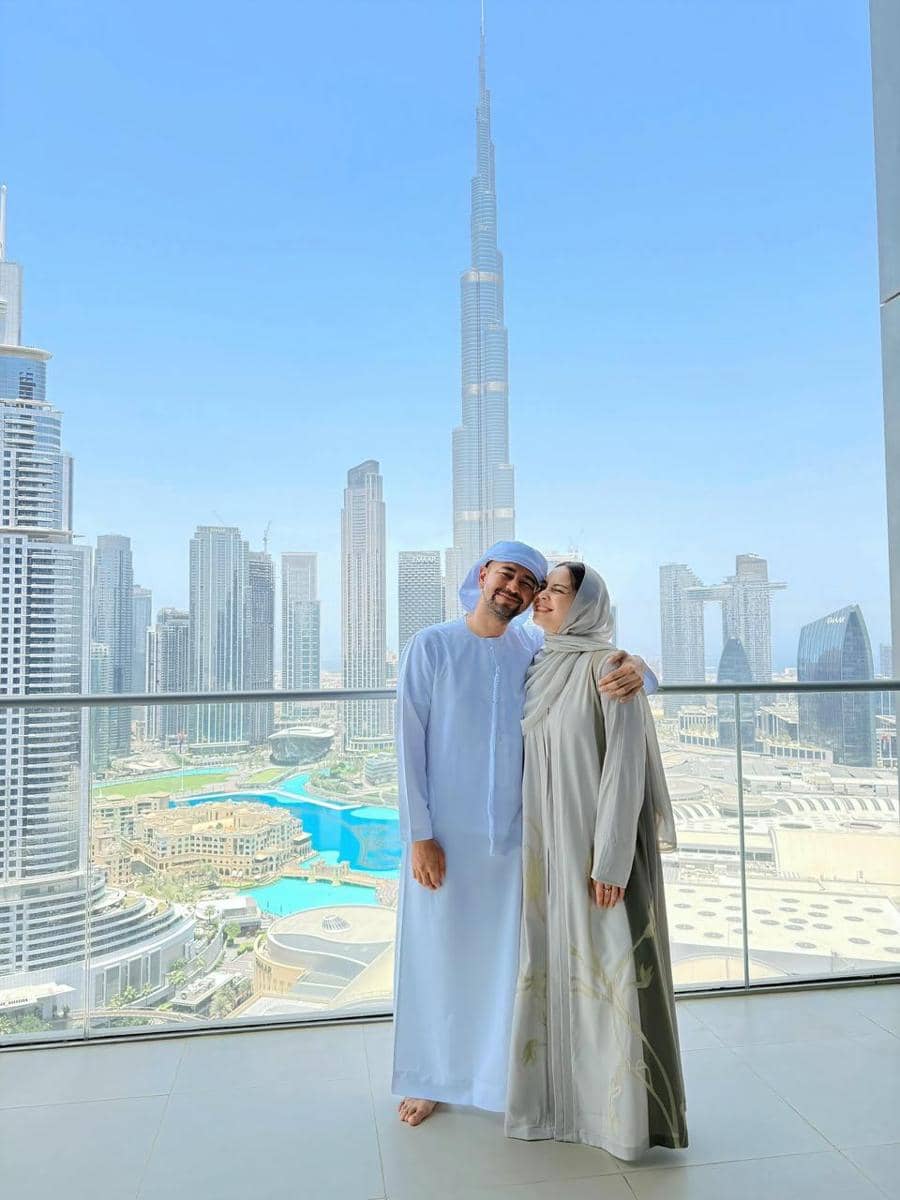 Sepasang wisatawan berpose di balkon dengan latar gedung pencakar langit Burj Khalifa dan panorama kota Dubai yang cerah.