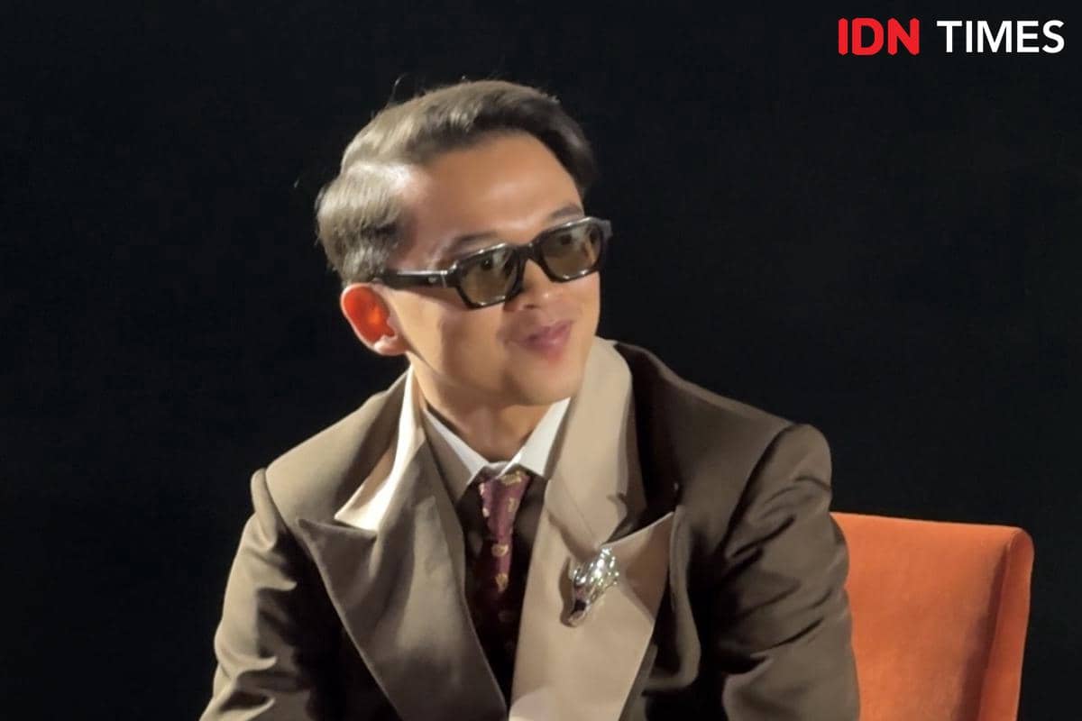 Seorang pria mengenakan setelan jas cokelat dan kacamata hitam duduk di kursi oranye saat konferensi pers film Monster Pabrik Rambut.