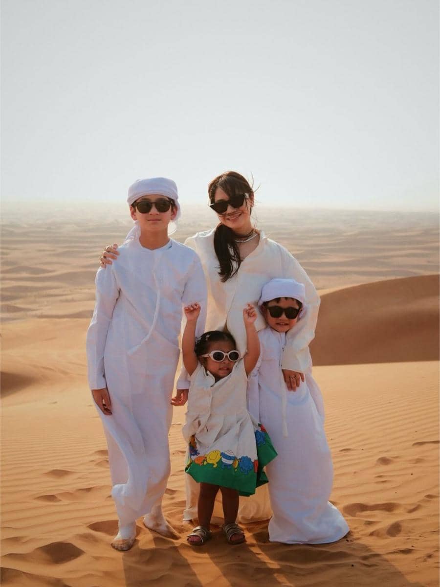 Seorang wanita bersama tiga anak mengenakan pakaian putih dan kacamata hitam berpose di padang pasir Dubai saat liburan.