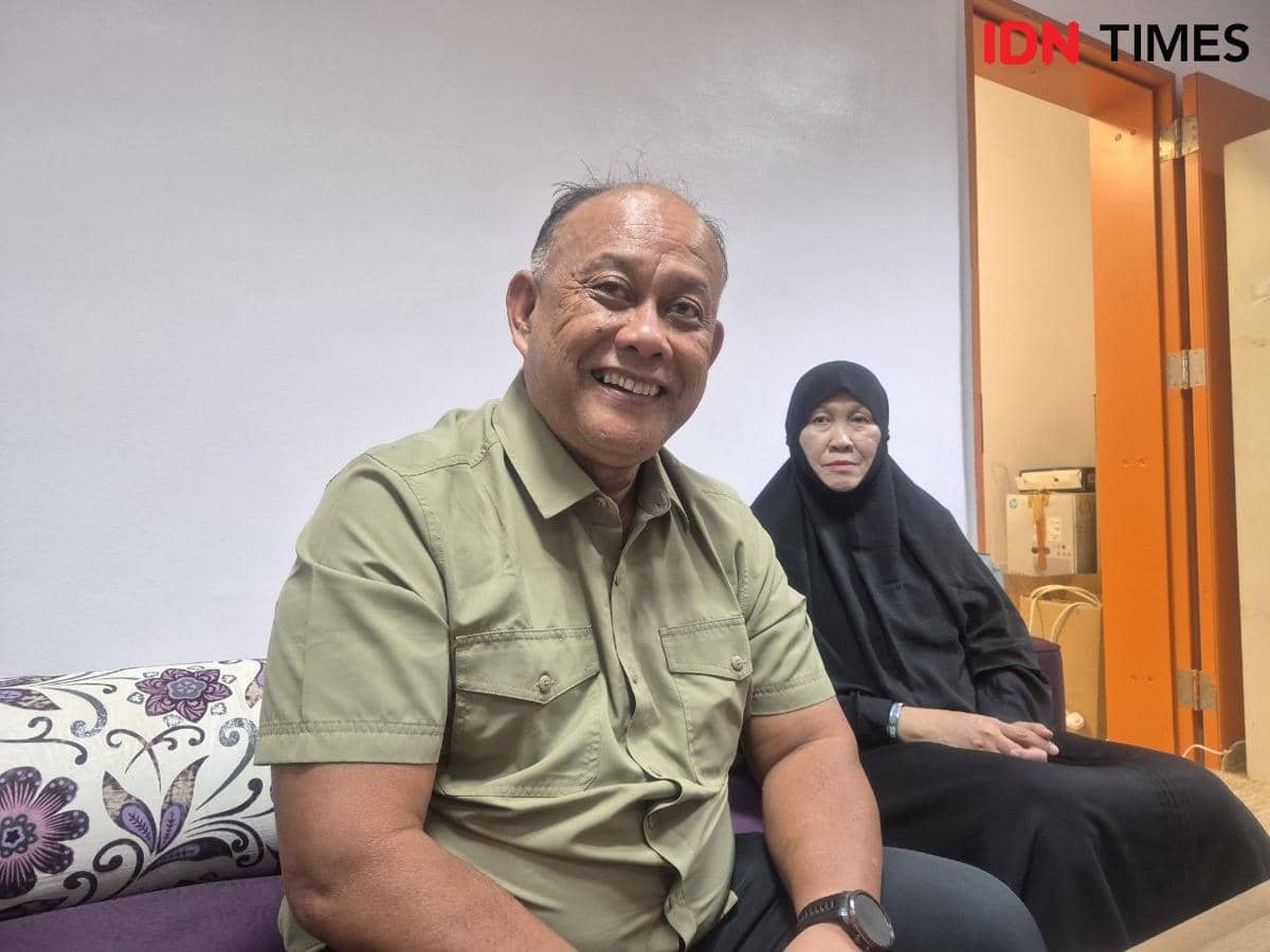 Seorang pria tersenyum duduk di sofa bersama seorang wanita berhijab hitam di ruangan dengan pintu oranye terbuka di belakang mereka.
