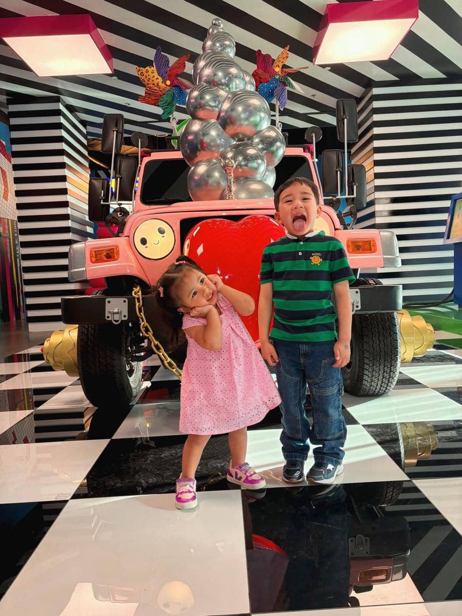 Dua anak kecil berpose ceria di depan mobil hias berwarna pink dengan dekorasi balon perak dan latar berpola hitam putih di Dubai.