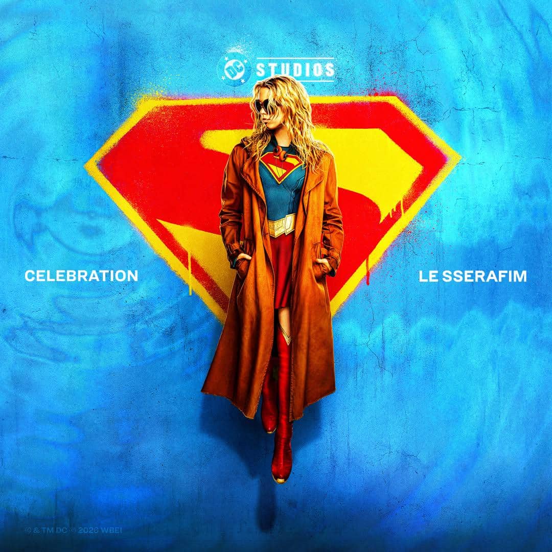 Seorang perempuan mengenakan kostum Supergirl dengan mantel cokelat berdiri di depan logo besar huruf S berwarna merah dan kuning di latar biru.