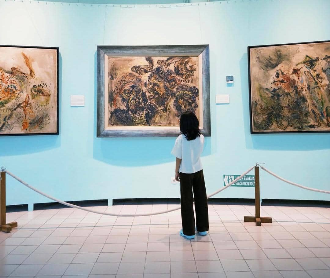 Seorang anak berdiri di depan tiga lukisan abstrak besar yang dipajang di dinding biru muda di sebuah galeri seni di Yogyakarta.