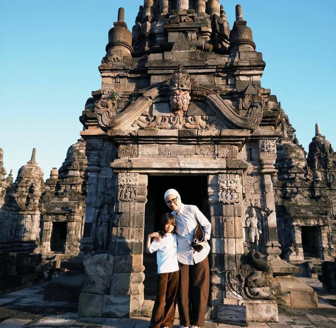 Seorang perempuan dan anak berpose di depan candi batu kuno di Yogyakarta dengan langit cerah di latar belakang.