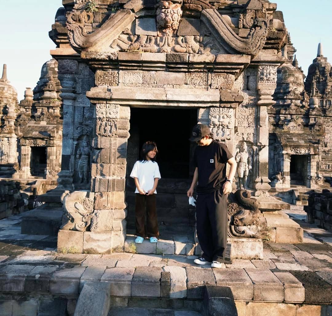 Dua orang berdiri di depan bangunan candi batu kuno di Yogyakarta dengan ukiran detail pada dinding dan suasana sore hari.
