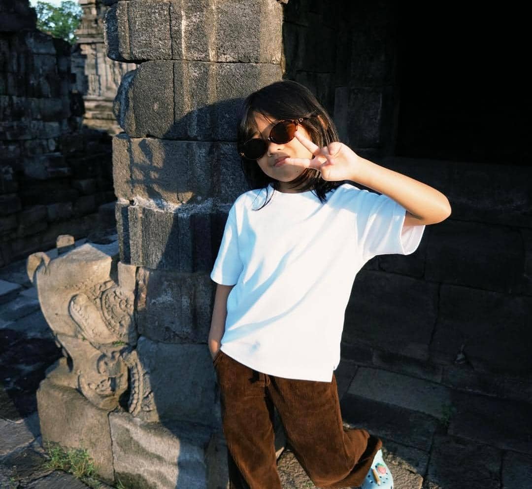 Seorang anak mengenakan kacamata hitam dan kaus putih berpose dengan tanda damai di area candi batu di Yogyakarta.