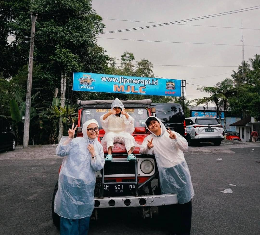 Tiga orang mengenakan jas hujan berpose di depan jip wisata Merapi di Yogyakarta dengan latar pepohonan dan papan jipmerapi.id.