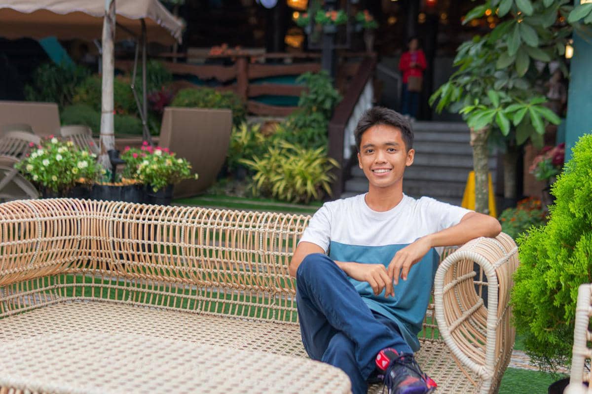 Seorang pria muda duduk santai di kursi rotan luar ruangan dengan latar taman hijau dan tangga menuju bangunan di belakangnya.