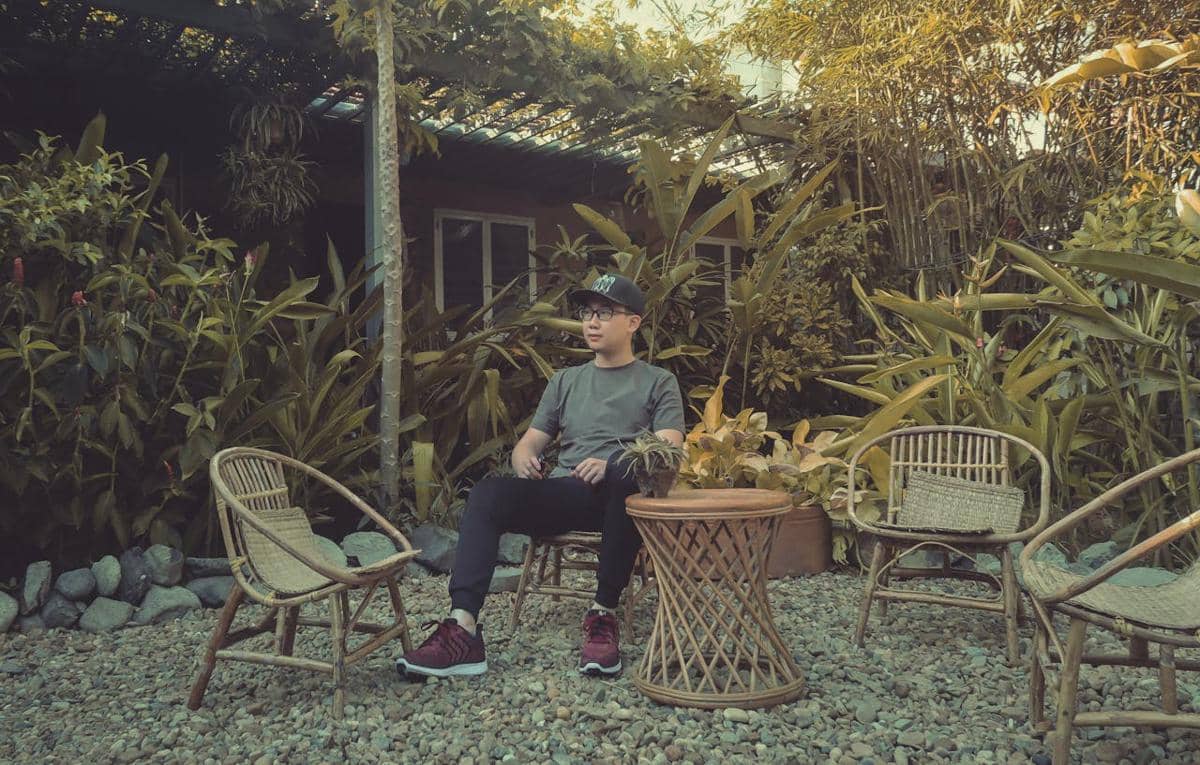 Seorang pria duduk santai di kursi rotan di taman dengan tanaman hijau lebat di sekelilingnya pada sore hari yang tenang.