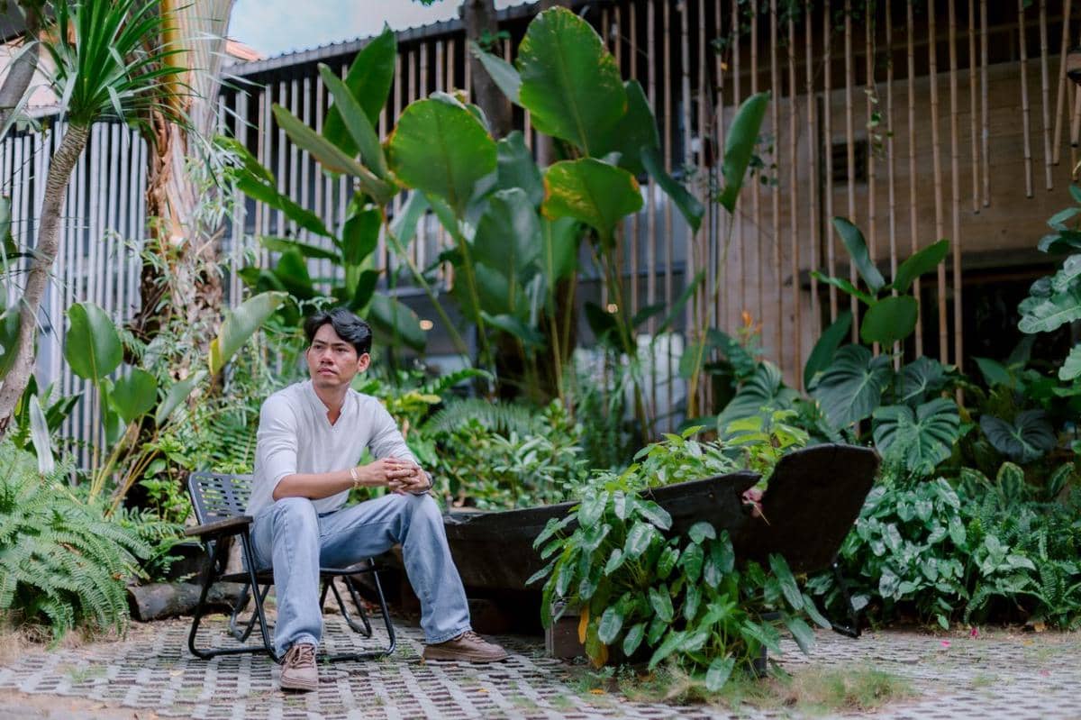 Seorang pria duduk sendirian di kursi taman dikelilingi tanaman hijau tropis dengan latar bangunan kayu dan bambu.