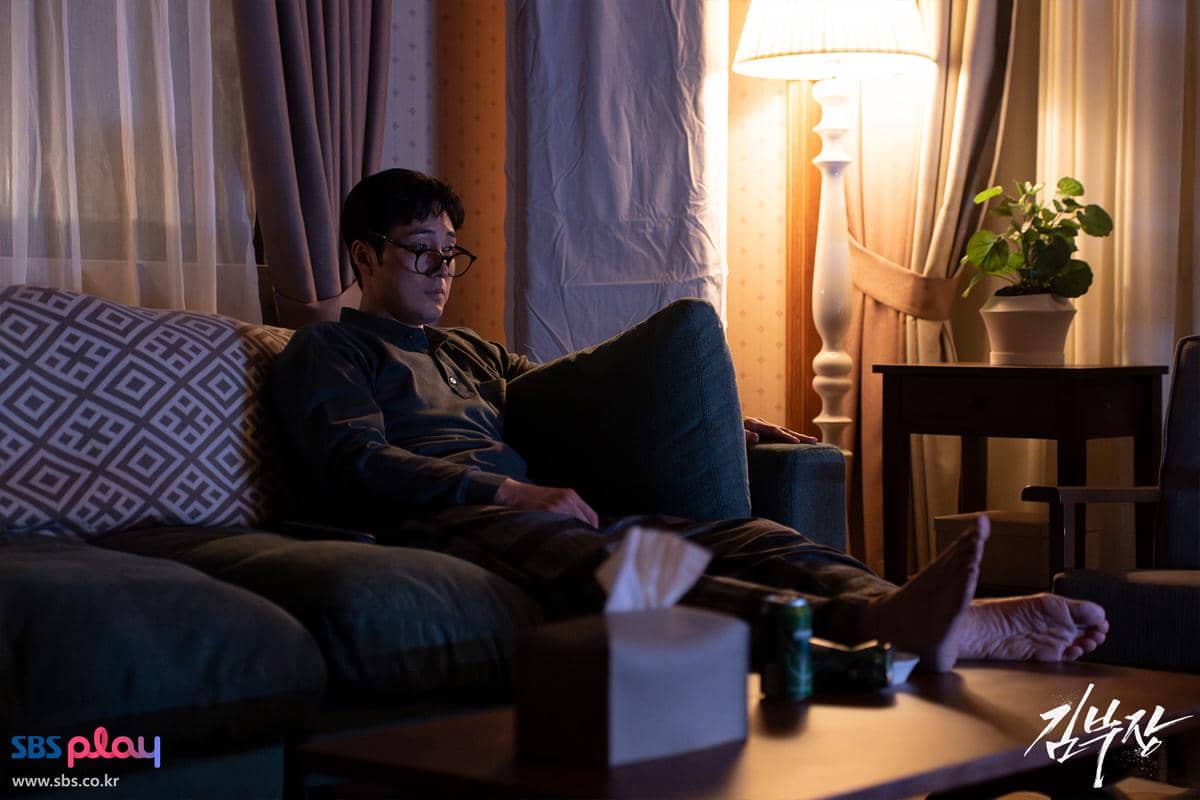 Seorang pria duduk di sofa ruang tamu dengan pencahayaan lampu hangat, mengenakan kacamata dan pakaian santai di malam hari.