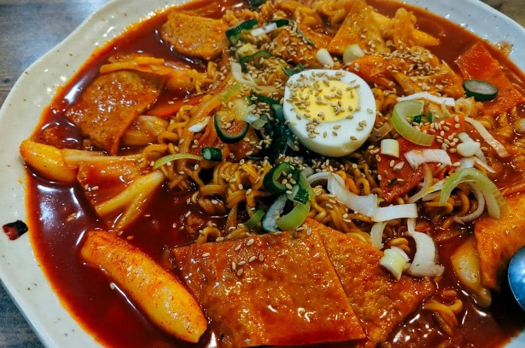 Semangkuk topokki dengan saus merah pedas, potongan kue beras, telur rebus, mi, irisan daun bawang, dan taburan wijen.