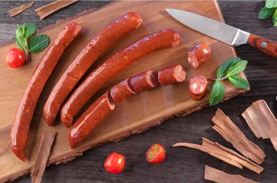 Beberapa potong sosis daging di atas talenan kayu dengan pisau, daun mint, dan tomat ceri sebagai hiasan di sekitarnya.