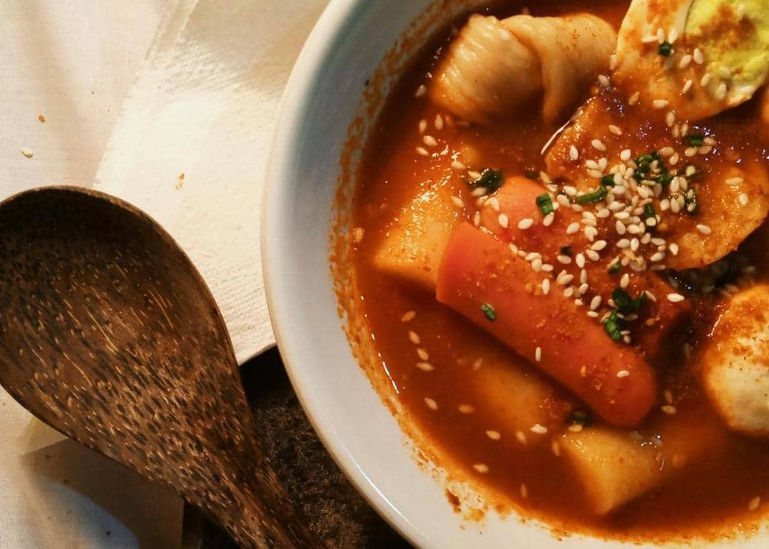 Semangkuk topokki berkuah merah pedas dengan taburan biji wijen dan daun bawang, disajikan di atas meja dengan sendok kayu.