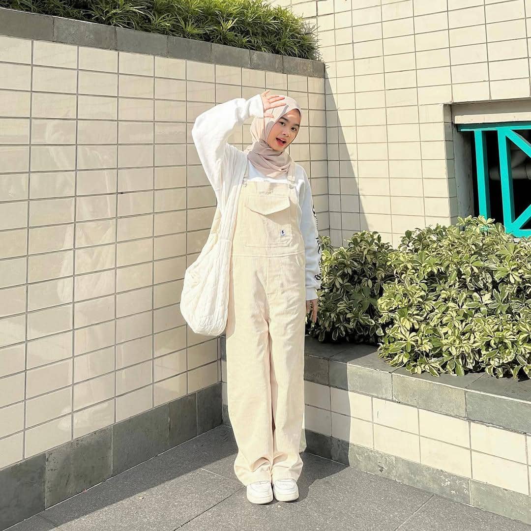 Remaja berhijab mengenakan overall krem dan sweater putih berpose di luar ruangan dengan latar dinding keramik dan tanaman hijau.