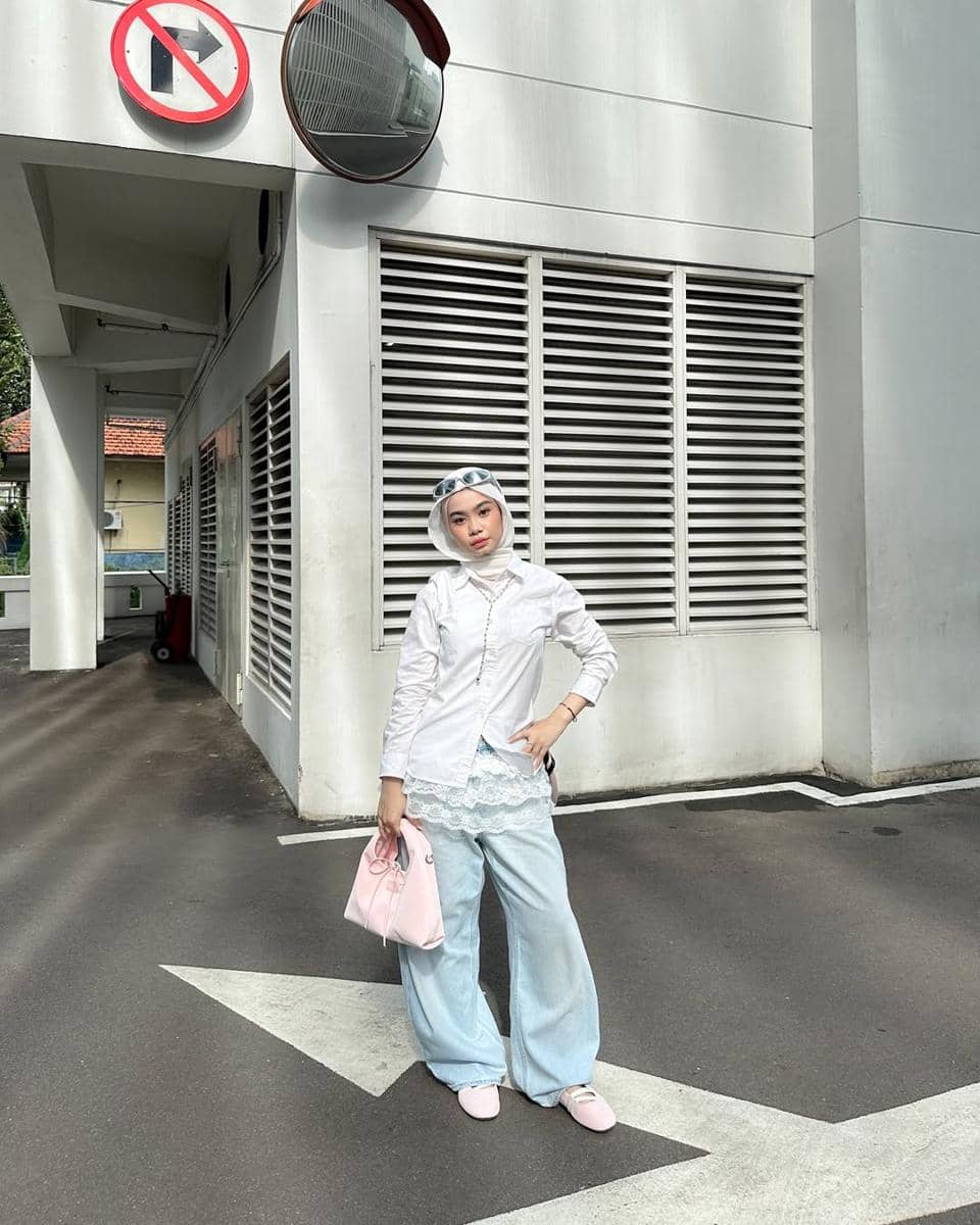 Seorang remaja berhijab bergaya kasual mengenakan kemeja putih, celana jeans longgar, dan tas pink di area parkir dengan dinding putih.