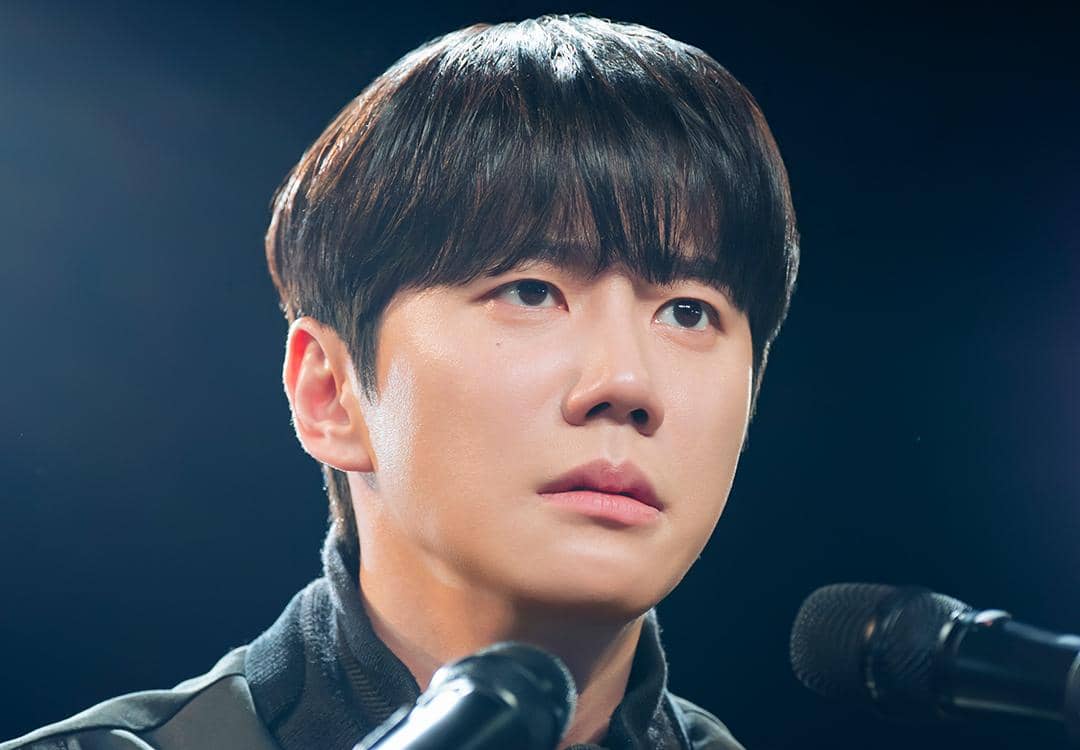 Seorang pria muda berambut hitam menatap serius di depan dua mikrofon dalam adegan drama Korea Reborn Rookie.