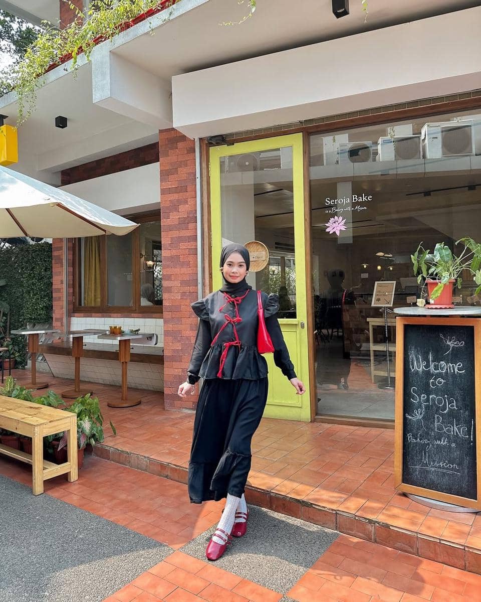 Seorang remaja berhijab mengenakan outfit kasual berwarna hitam dengan aksen merah dan tas merah, berpose di depan kafe Seroja Bake.