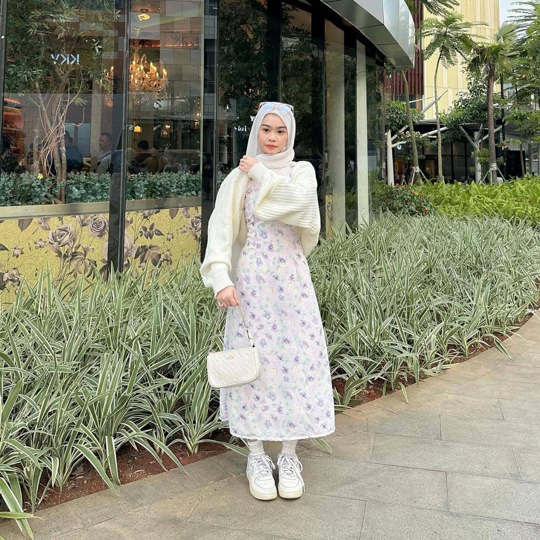 Seorang remaja berhijab mengenakan dress floral ungu muda, kardigan putih, dan sepatu sneakers putih di area luar gedung modern.