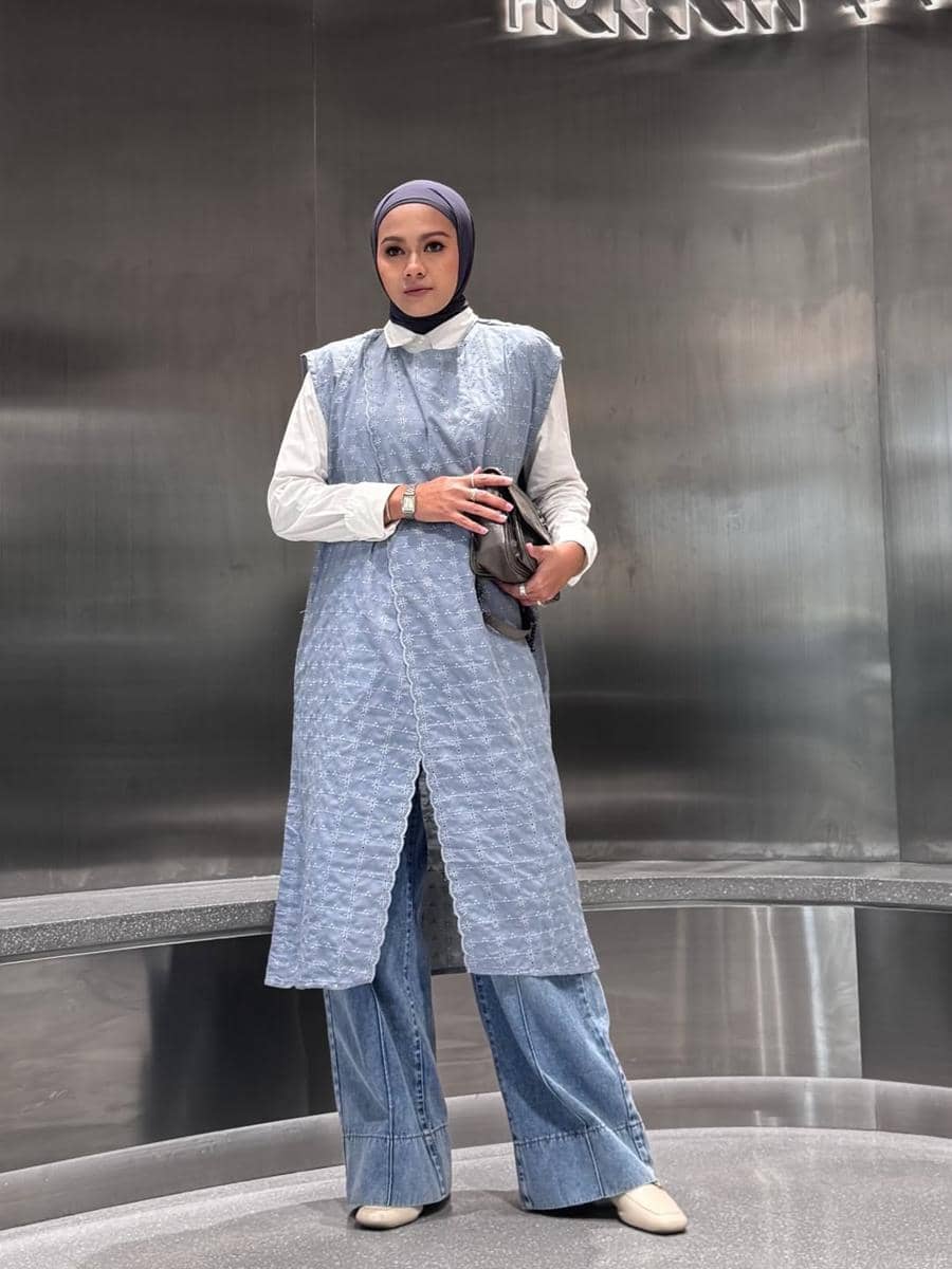 Seorang wanita berhijab mengenakan kemeja putih, outer biru bermotif, dan celana jeans lebar sambil memegang tas tangan di ruangan modern.