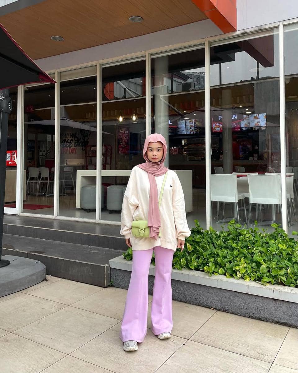 Remaja berhijab mengenakan sweater krem, celana ungu muda, dan tas kecil hijau muda berdiri di depan restoran modern.
