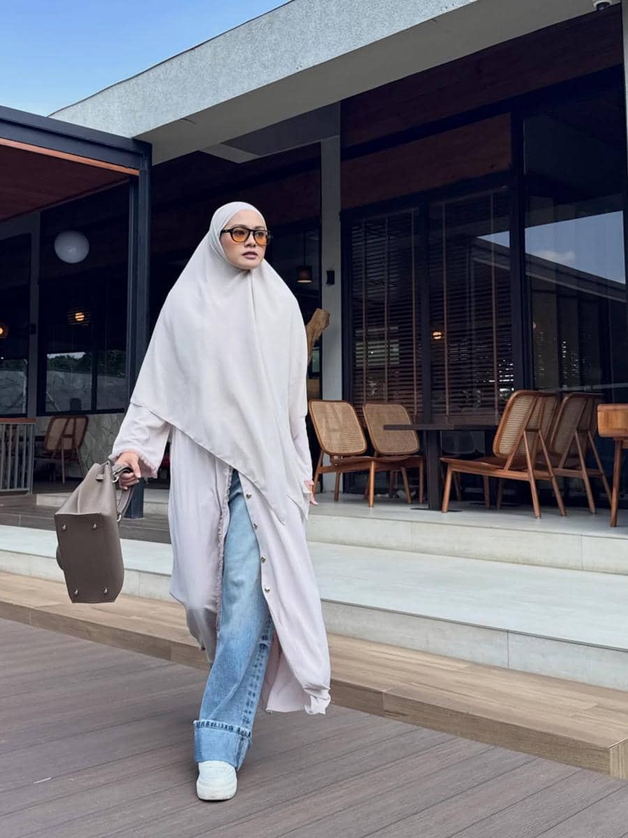 Seorang wanita berhijab panjang berwarna abu-abu muda mengenakan celana jeans longgar dan membawa tas abu-abu di area luar kafe modern.
