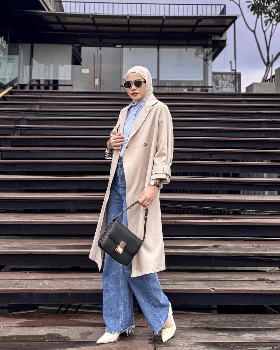 Seorang wanita berhijab mengenakan kemeja biru, celana jeans longgar, dan mantel krem sambil membawa tas hitam di tangga kayu.