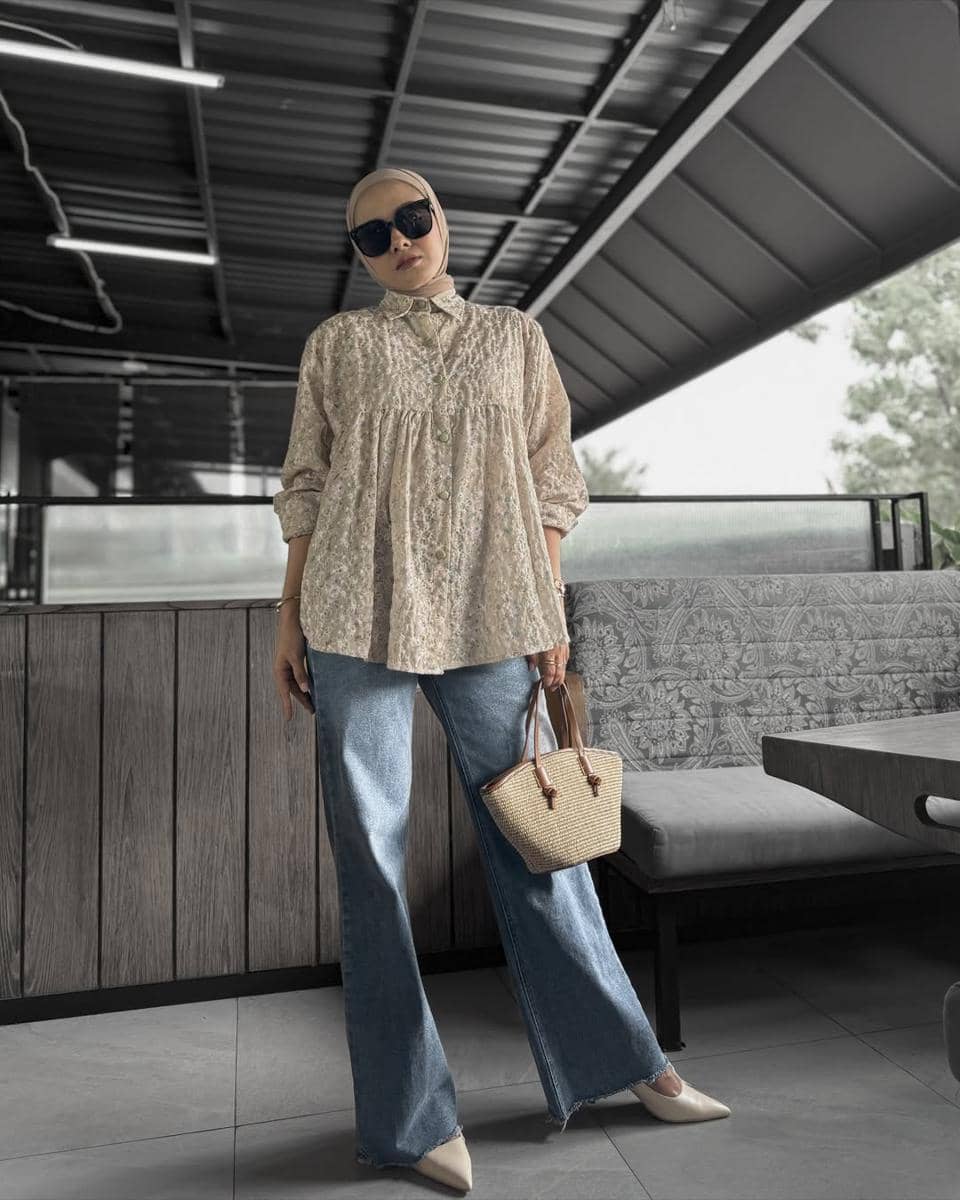 Seorang wanita berhijab mengenakan blus motif bunga dan celana jeans lebar, membawa tas rotan kecil serta memakai kacamata hitam.