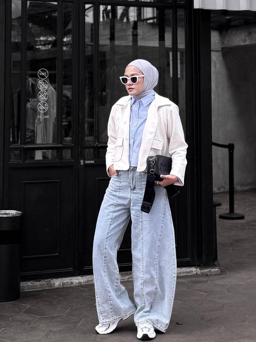 Seorang wanita berhijab mengenakan kemeja biru muda, jaket krem, celana jeans longgar, dan kacamata putih berdiri di depan pintu kaca hitam.