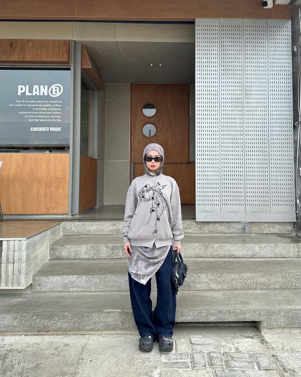 Seorang remaja berhijab bergaya kasual mengenakan sweater abu-abu bermotif, celana longgar gelap, dan kacamata hitam di depan kafe modern.