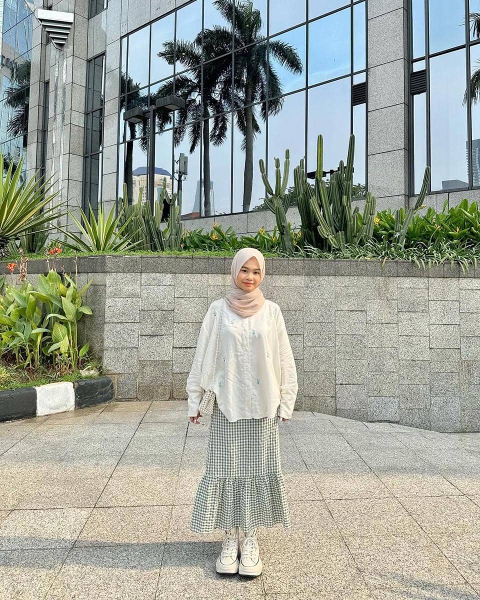 Seorang remaja berhijab mengenakan outfit kasual berupa blus putih longgar dan rok kotak-kotak hijau di depan gedung bertingkat kaca.