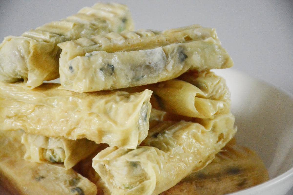 Tumpukan dimsum kulit tahu berwarna kuning keemasan dengan isian sayuran dan daging, disajikan di atas piring putih.