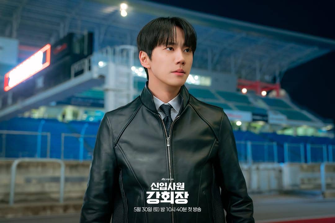 Seorang pria mengenakan jaket kulit hitam dan dasi berdiri di area stadion pada malam hari dalam cuplikan drama Reborn Rookie.