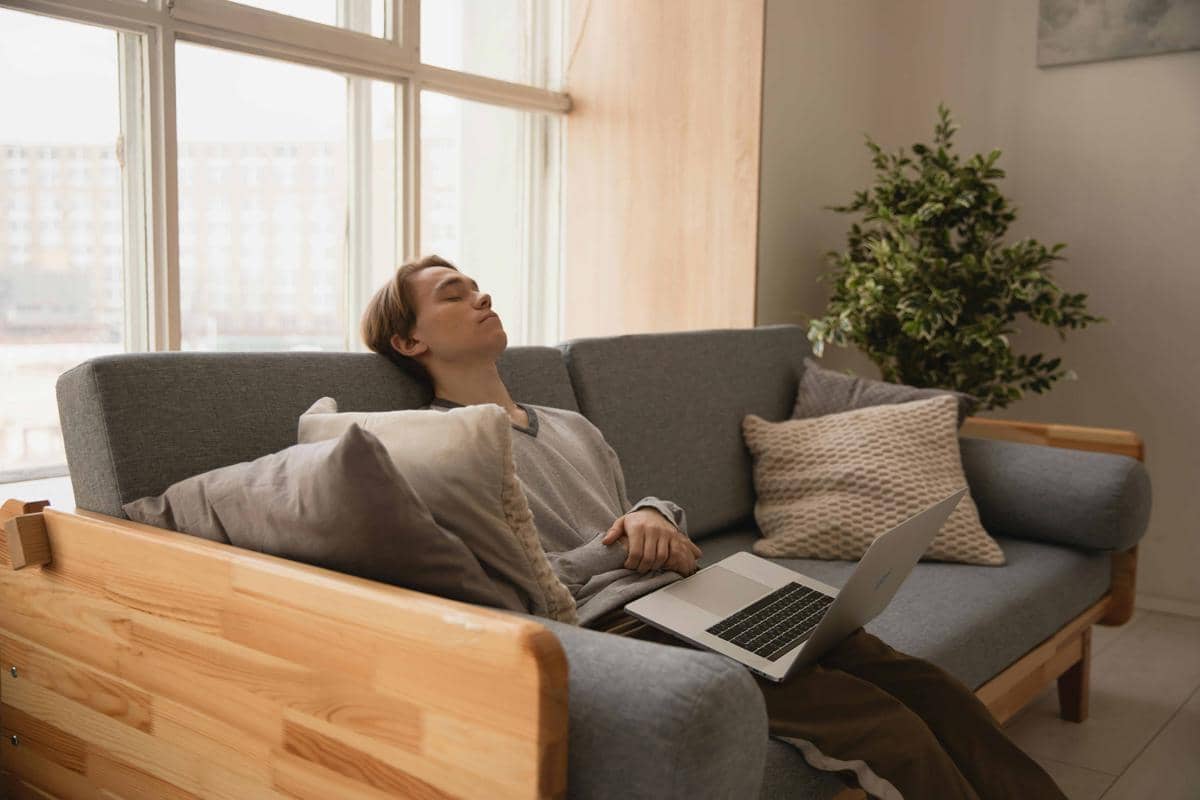 Seseorang duduk di sofa abu-abu dengan laptop di pangkuan, tampak lelah dan memejamkan mata di ruang tamu yang terang.