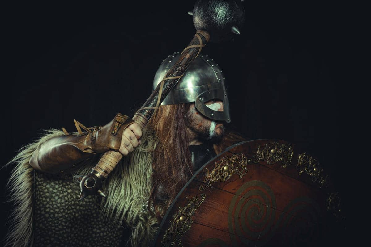 Pria berpakaian prajurit Viking mengenakan helm besi sambil memegang senjata mace berduri dan perisai kayu dengan latar belakang hitam.