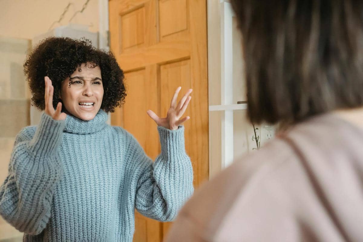 Seorang perempuan mengenakan sweter biru sedang marah dan mengangkat tangan sambil berbicara kepada seseorang di depannya.