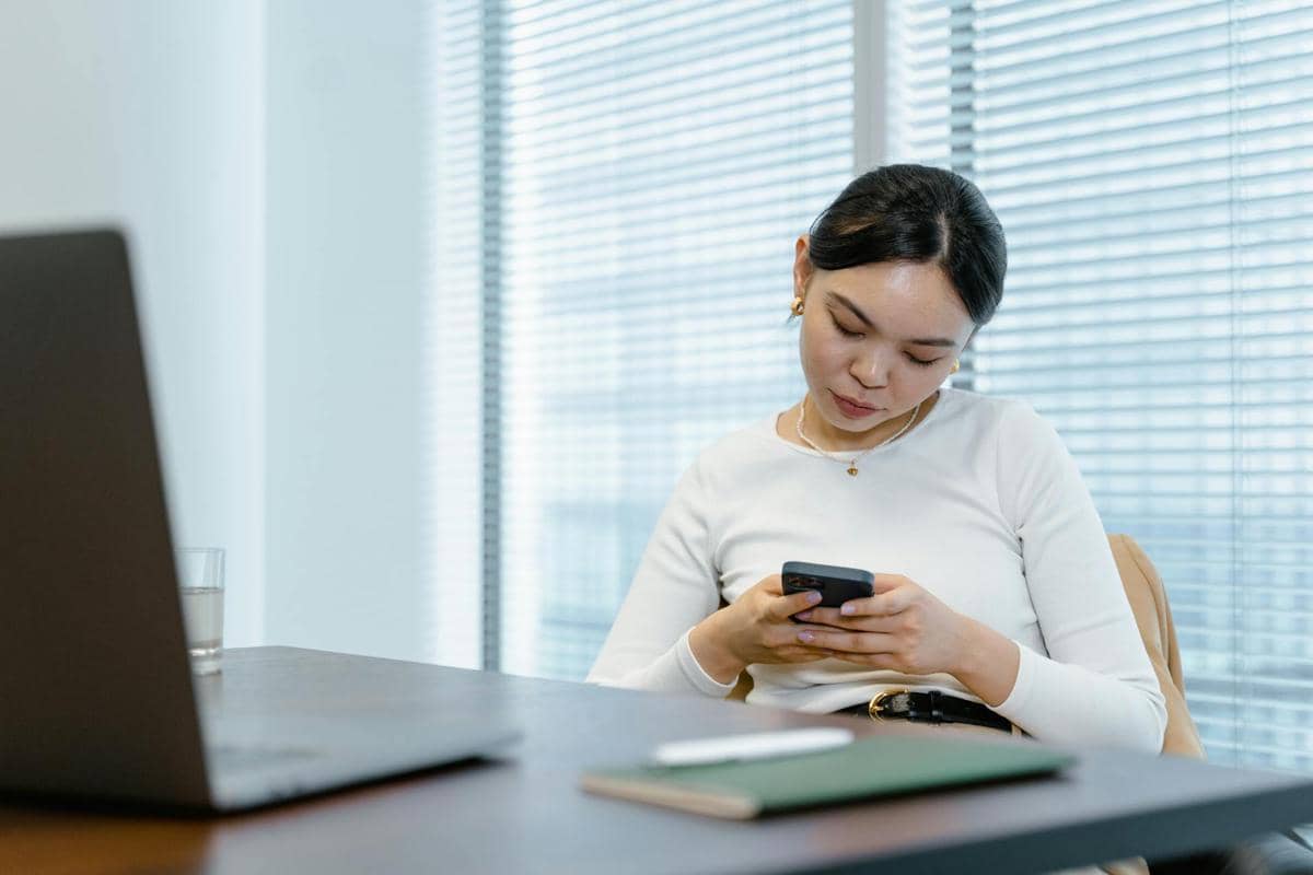 Seorang wanita duduk di meja kantor sambil melihat ponsel, dengan laptop dan buku catatan di depannya di ruangan bercahaya alami.